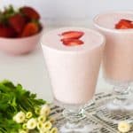 Strawberry Lassi in 2 glasses with flowers on the left and strawberries at the back
