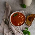 Roasted red pepper and tomato soup in a white bowl with a spoon on the left and grilled cheese sandwich on the right
