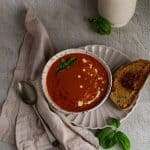 Roasted red pepper and tomato soup in a white bowl with a spoon on the left and grilled cheese sandwich on the right